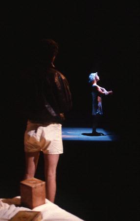 A person in a dark room stands in their underwear with a leather jacket, facing another person performing in a spotlight. The performer appears to be in motion, wearing a dress, with arms raised. A box is in the foreground.