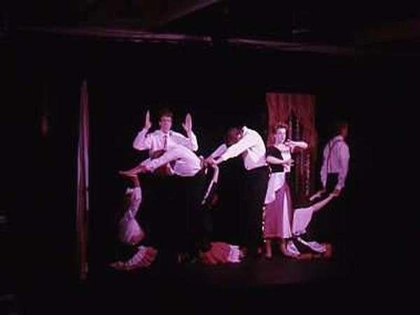 A group of performers on stage create various poses forming letters or shapes. They are in semi-formal attire against a dark background with a curtain. The lighting highlights them, leaving the rest of the stage in shadow.