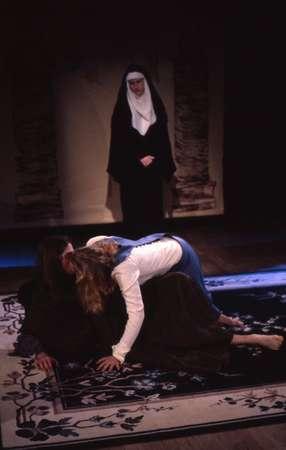 A person in a nun's habit watches as two people lie on a patterned rug on stage; one person leans over the other. The background is dimly lit, creating a dramatic atmosphere.