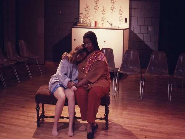 Two women sit on a bench in a dimly lit room. One woman in a gray sweater and shorts leans on the other, who wears a colorful top and orange pants. They are surrounded by empty chairs on a wooden floor, with a small kitchen area in the background.