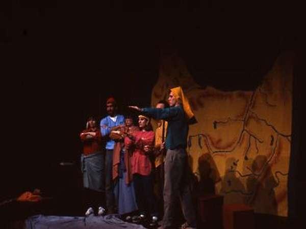 A group of actors on stage performs a play. They stand in front of a backdrop that resembles an ancient map. One actor is gesturing forward dramatically while others watch attentively. The lighting is dim, casting shadows across the stage.