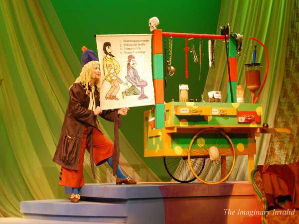 A performer in a curly wig and costume points at a colorful cart displaying anatomical diagrams and medical tools on stage. The background is green with flowing curtains. The setup suggests a theatrical or comedic performance related to medicine.