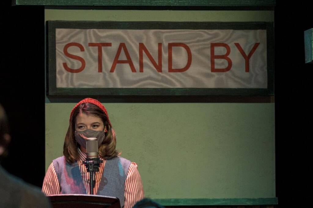 A person wearing a red headband and a face mask stands in front of a microphone, positioned beneath a large sign that reads "STAND BY." The background is dark, suggesting a stage or studio setting.