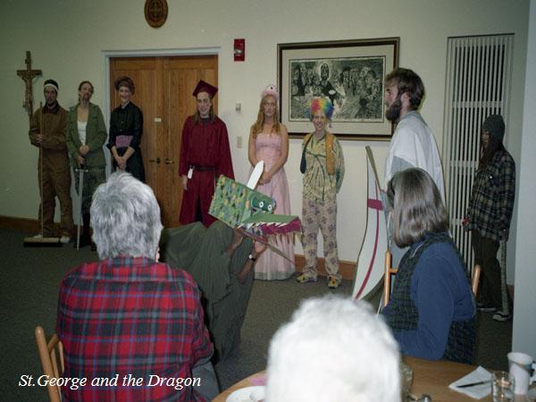 A group of people in costume perform a skit called "St. George and the Dragon." The scene features a person in a dragon costume and others dressed as various characters. Two audience members are seated in the foreground, watching.