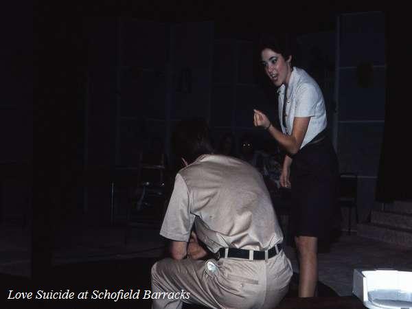 A woman in a dark dress gestures passionately toward a seated man in a uniform, on stage during a performance titled "Love Suicide at Schofield Barracks". The background is dimly lit, suggesting a theatrical setting.