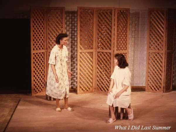 Two actors perform a scene on stage from "What I Did Last Summer." One is standing, gesturing with her hands, while the other sits on a chair. They are both wearing loose, light-colored clothing. Wooden lattice panels form the backdrop.