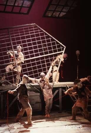 Theatrical scene from "Macbeth" featuring actors in tattered costumes. They are dramatically interacting with a large, tilted metal grid frame on a stage, conveying a sense of struggle or conflict. The background is dimly lit.