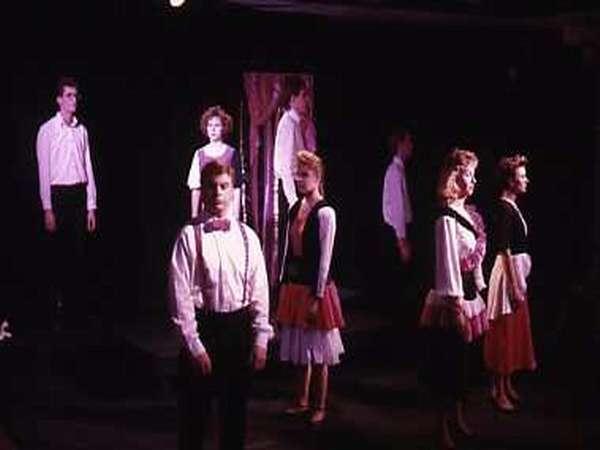 A group of performers stands on stage, with men in white shirts and women in colorful skirts. They are in a theatrical pose, with dim lighting and a reflective backdrop adding to the dramatic atmosphere.