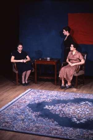 Three people in a room with a patterned rug. Two are seated at a wooden table, and one stands behind them. The seated individuals are dressed in dark and patterned attire, with the standing figure wearing black. The background is dark with a red square.