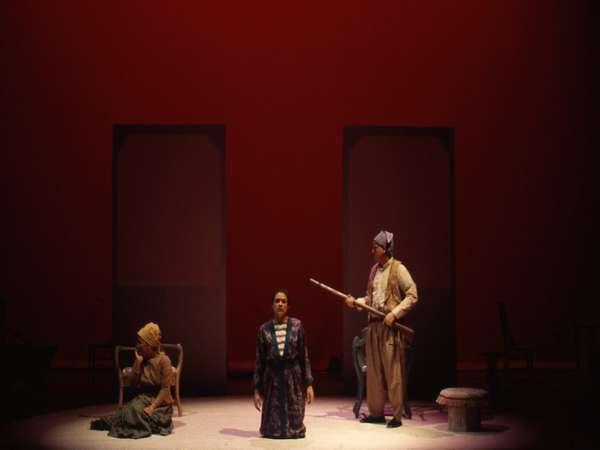 Three actors perform on a dimly lit stage with a red background. One is kneeling in the center, another stands holding a long stick, and the third sits on the floor. There is minimal scenery, creating a dramatic atmosphere.