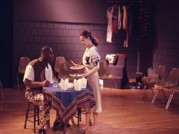 Two people perform a scene on stage. One, seated at a table with a blue cloth and white takeout boxes, looks at the standing person who holds a tray. The background shows chairs and hanging clothes, suggesting a rehearsal or casual theater setting.