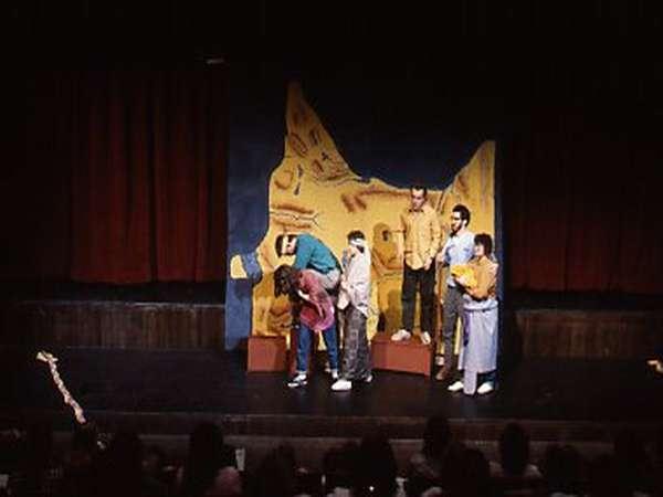 A group of actors performs on stage in front of an audience. The backdrop features a large, abstract yellow design. The actors are wearing casual clothes and are engaged in a dynamic scene.