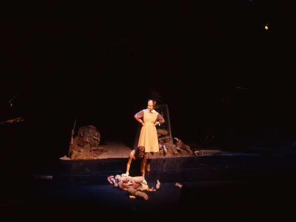 A dimly lit stage scene shows a person in a dress standing with hands on hips, looking down at another person lying on the floor. The background features scattered props suggesting a dramatic or theatrical setting.