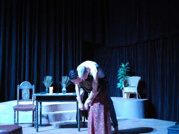 Two actors perform on a dimly lit stage. One person leans over the other in a dramatic pose. A table, chairs, and plants are part of the set. Long curtains and a spotlight create a theatrical atmosphere.