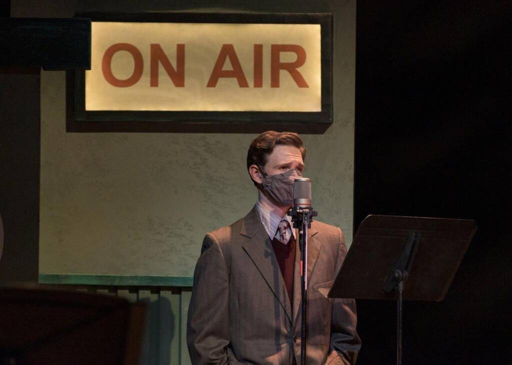 A person wearing a suit and face mask stands in front of a vintage microphone, reading from a script. A sign reading "ON AIR" is illuminated in the background, suggesting a radio broadcast setting.