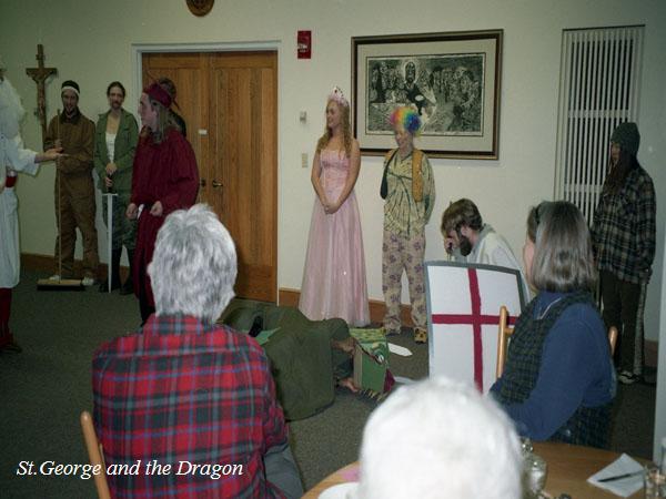 Several people dressed in costumes are in a room. One person is in a suit of armor, others are wearing a gown, a clown costume, and various medieval attire. An audience is seated nearby, watching the performance. A poster with "St. George and the Dragon" is visible.