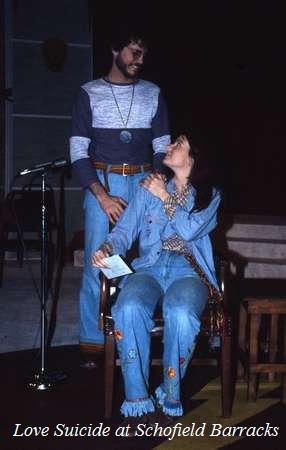 A man stands smiling beside a seated woman, who looks up at him. Both are wearing denim outfits. A microphone stand is visible in the background. The text "Love Suicide at Schofield Barracks" is at the bottom.