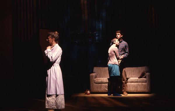 Three people on stage in dim lighting. Two stand close together near a couch, engaged in conversation. A third person stands apart, facing away, with a contemplative expression. The setting suggests a dramatic scene.