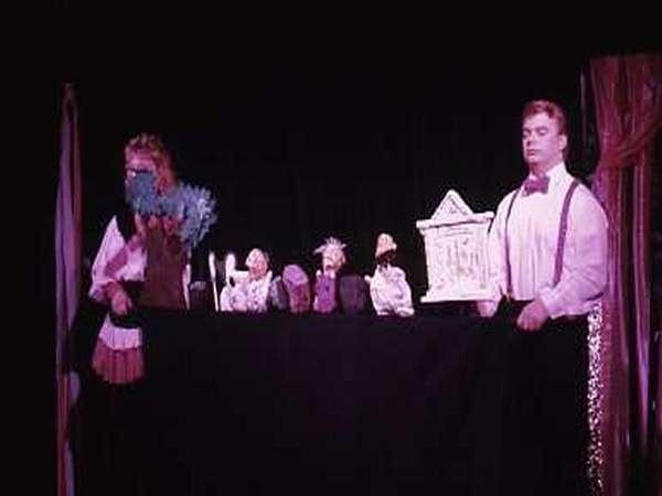 Two performers in costume stand on either side of a black table with various puppets and a small theater backdrop on it. The scene is dimly lit, suggesting a theatrical performance setting.