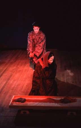 Two performers in traditional Japanese attire stand and kneel on a dimly lit wooden stage. One is kneeling beside a rectangular wooden frame. The scene is illuminated with a soft, warm light creating a dramatic atmosphere.