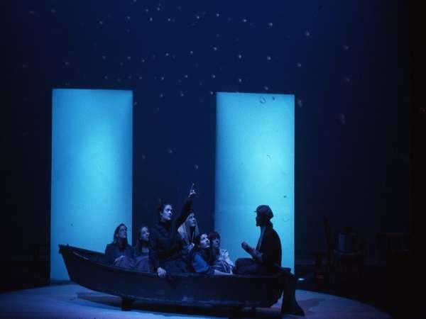 A theatrical scene featuring a group of people in a boat on stage, with two large illuminated panels behind them. The background is dark, resembling a night sky with stars. One person points upwards, creating a sense of wonder or storytelling.