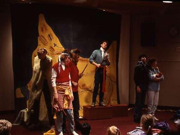 A group of people performs a theatrical scene on stage. Some are standing on a platform, while others are on the stage floor. They are dressed in casual clothes and props. A painted backdrop is visible behind them, depicting an abstract scene.