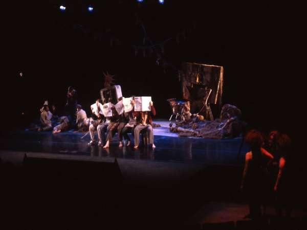 A dimly lit stage with people sitting in a row, some holding large rectangular objects. Others are scattered around the stage in various poses, with rocky props and a dark backdrop adding a dramatic effect.