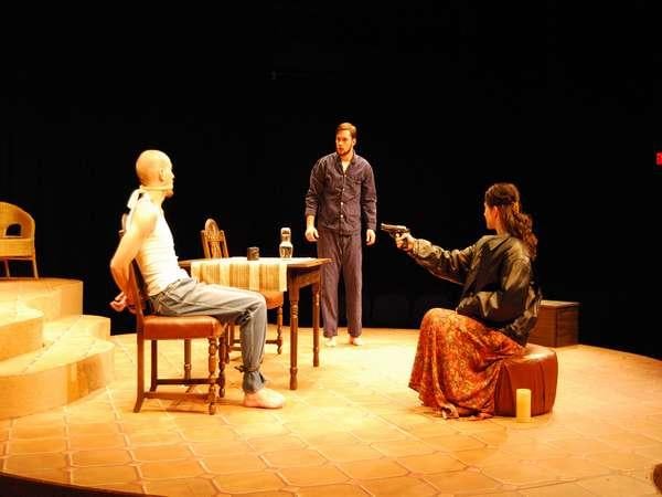 Three people are on stage in a dramatic scene. One person is seated with their arms behind the chair, another stands nearby, and the third, seated on the floor, points a gun at the seated person. The stage is dimly lit with a table and chairs.