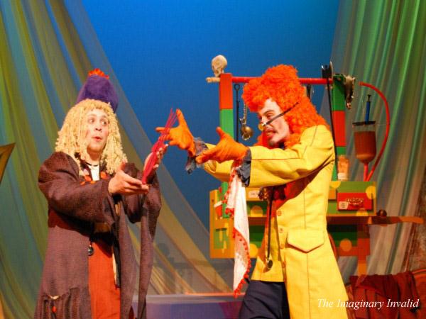 Two actors in colorful, eccentric costumes perform on stage. One wears a shaggy wig and holds a red book, while the other, in a bright yellow outfit and orange wig, gestures animatedly. The backdrop features vibrant props and drapery. Text reads "The Imaginary Invalid.