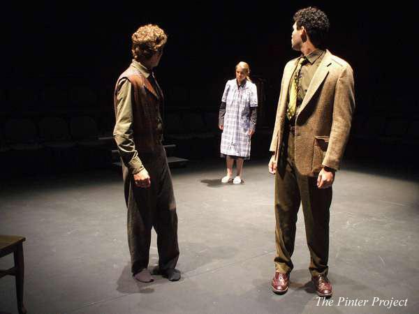 Three actors on a dimly lit stage perform a scene. Two men in suits stand facing a woman in a plaid dress and slippers. Chairs are arranged in a circle around the stage. The words "The Pinter Project" are on the bottom right.