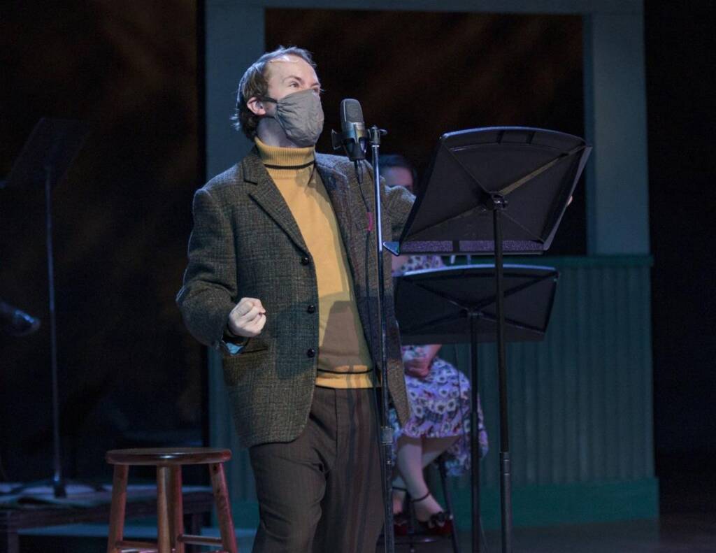 A person wearing a mask and a tweed jacket stands at a microphone, holding a script. They are in a recording studio or theater setting, with a stool and another person visible in the background.