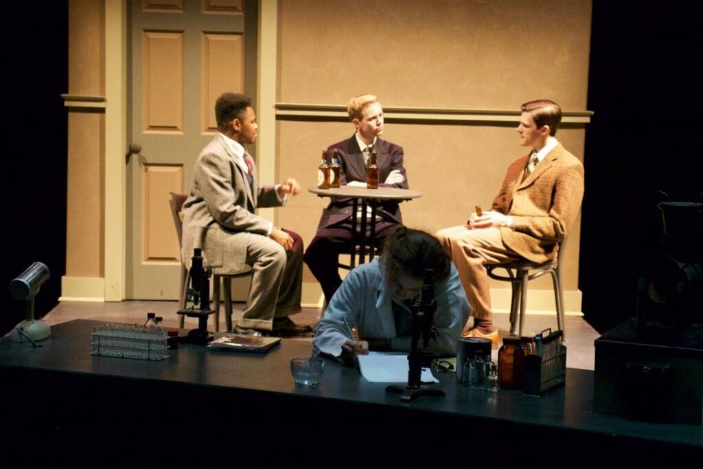 A theatrical scene depicts three men in suits sitting at a round table, engaged in conversation, with another person in a lab coat examining something under a microscope at a desk in the foreground. A closed door and dim lighting set the background.