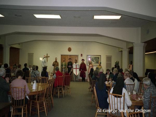 A theatrical performance takes place in a large room. Actors are on a stage dressed in medieval-style costumes. An audience sits at tables watching the play. The text "St. George and the Dragon" is visible in the bottom right of the image.