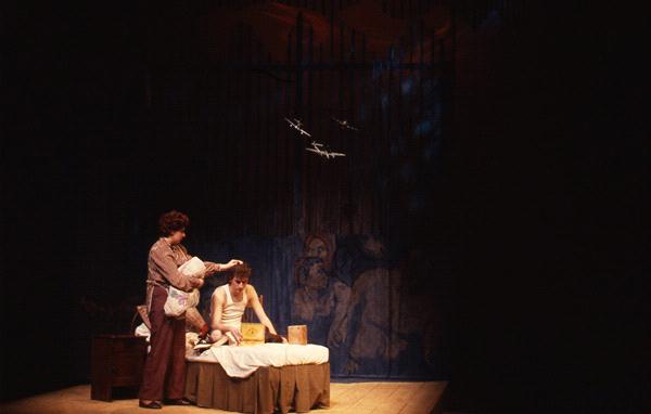 A dimly lit stage with two actors. One actor, seated on a bed, looks at a small box. The other, standing beside, offers a towel. The backdrop features a dark mural with faint images of people. Airplanes are projected on the wall above.