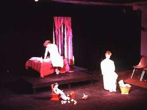 A dimly lit stage with two actors. One stands by a bed covered with a red blanket, and the other sits on a chair. A doll and toys are scattered on the floor near a basket. A window with pink curtains hangs in the background.