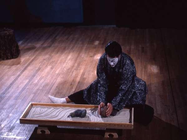 A person in traditional attire sits cross-legged on a wooden floor, facing a rectangular sand tray with a stone and a small rake. The person has a painted face and is holding something in their hands, appearing focused on the tray.