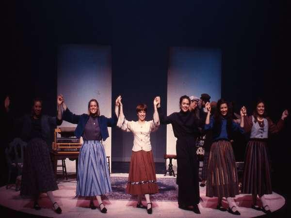 A group of six people on stage in a theater production, holding hands and raising their arms in celebration. They are dressed in period costumes, with long skirts and blouses, standing in a semicircle under stage lights.