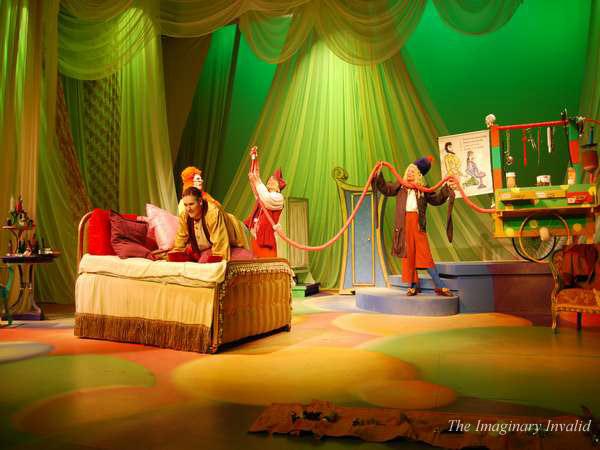 A vibrant stage scene from "The Imaginary Invalid" features actors in colorful costumes. One actor lies on a bed while others attend to them, surrounded by whimsical props and green drapery.