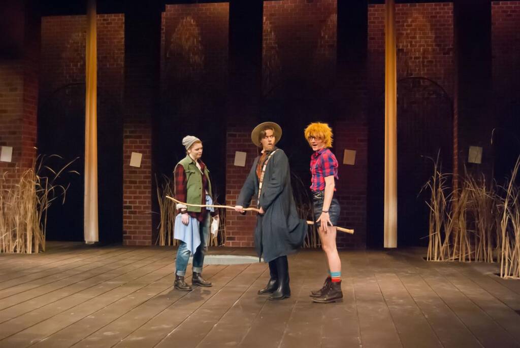 Three actors on a theater stage perform in front of a brick wall backdrop with tall, dried plants. One wears a beanie and green vest, another a wide-brimmed hat and dark cloak, and the third a red wig and checkered shirt. They hold a wooden stick.