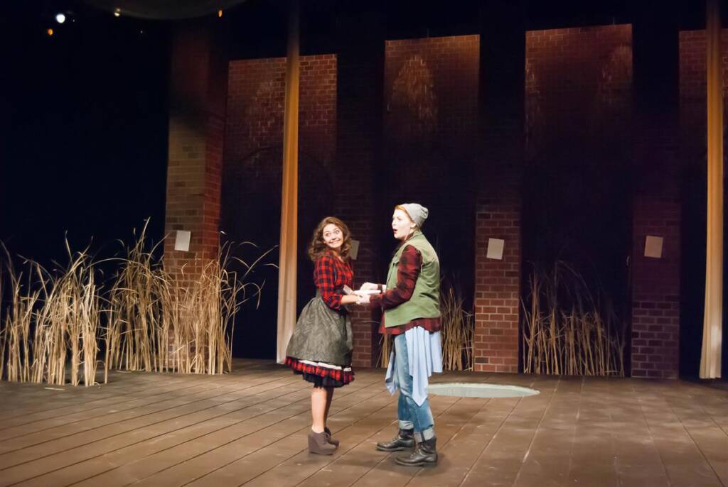 Two people in costume perform on a stage set resembling a rustic outdoor scene. One wears a plaid shirt and skirt, and the other a beanie, green jacket, and jeans. They face each other, holding hands, with tall grasses in the background.