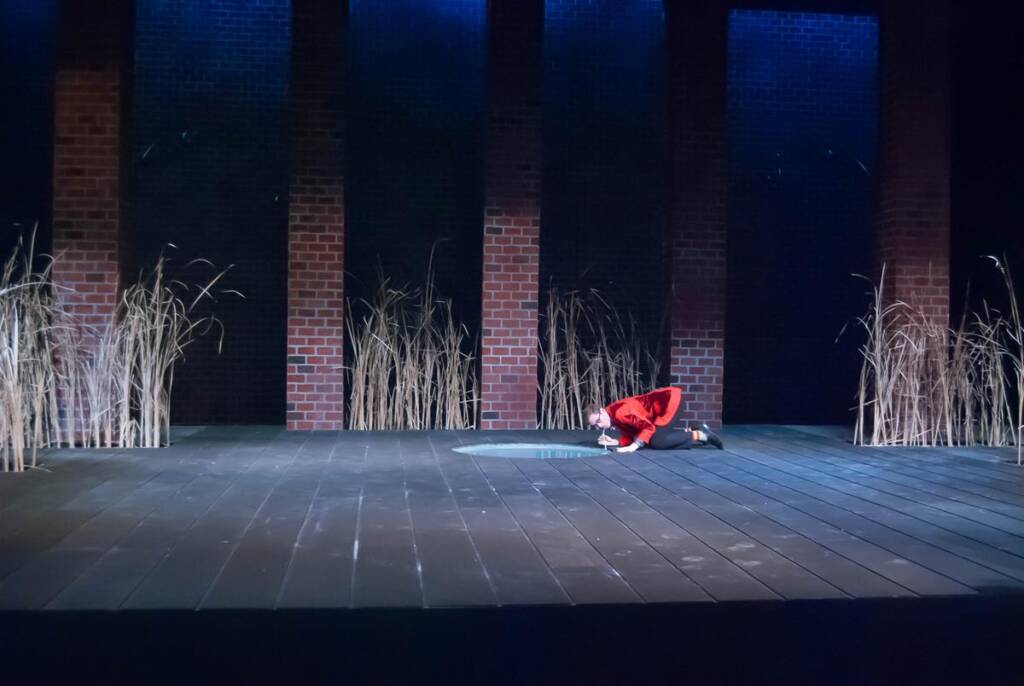 A person in a red outfit kneels on a large stage with a dark, wooden floor. Tall brown reeds are placed around brick columns in the background, with blue lighting illuminating the scene.