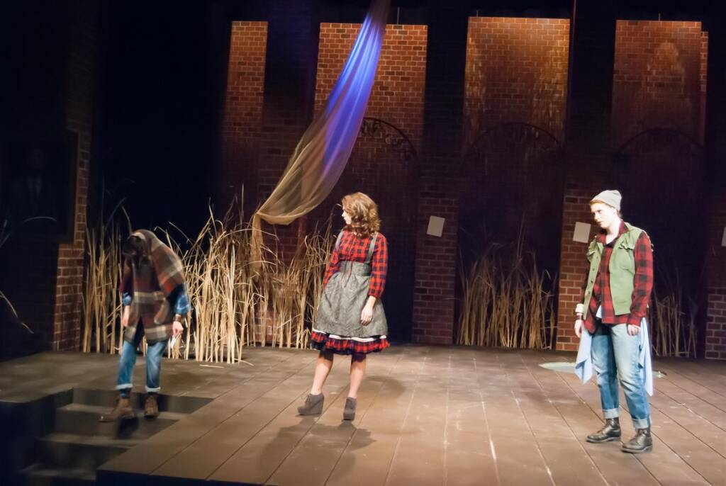 Three actors on stage perform in a rustic, theatrical setting with a backdrop of tall grasses and brick walls. One actor stands on steps, while the others wear plaid shirts and look toward them, creating a dramatic scene.