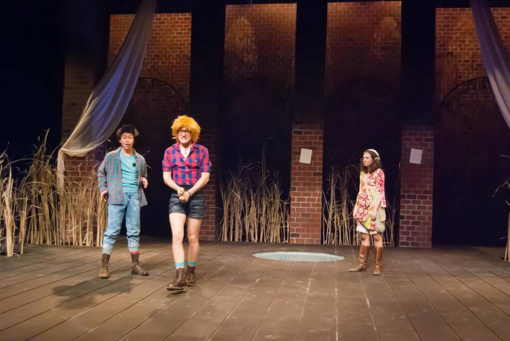 Three actors perform on a stage with a brick wall backdrop. One wears denim overalls, another wears a plaid shirt with shorts and a wig, and the third wears a floral dress. The floor is wooden with tall grass props. Curtains drape from above.