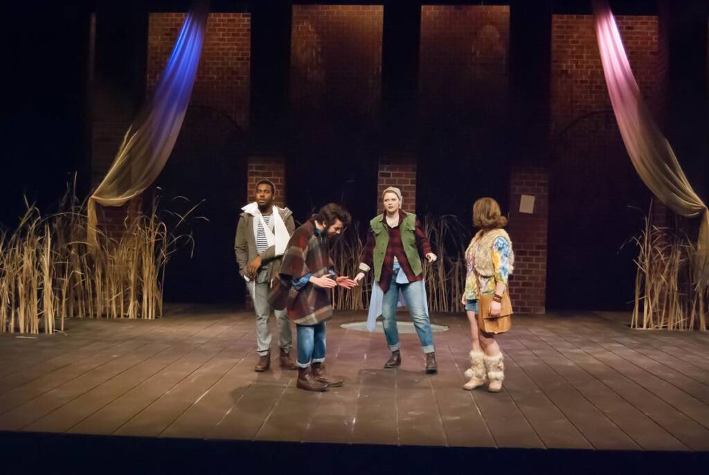 Four actors perform on a dimly lit stage with a rustic backdrop. They are surrounded by tall, dry grasses and draped fabric on brick walls. Each actor is dressed in casual, layered clothing, suggesting a contemporary or rustic setting.