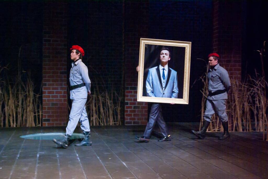 Three people in military-style uniforms walk across a stage. The central figure carries a large framed portrait of a person in a suit. The scene is set against a backdrop of brick walls and dry grass.