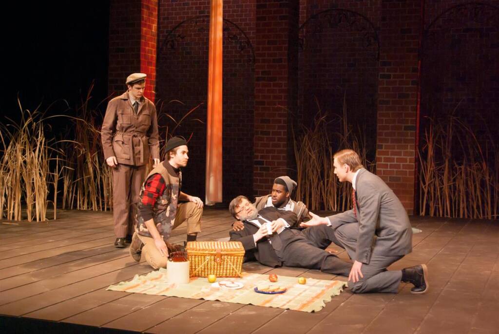 Five actors perform a stage scene. One is in military attire, another in flannel and boots, and three in suits. They are gathered around a picnic setup with a checkered cloth, basket, and scattered apples. The backdrop features tall reeds and brick arches.