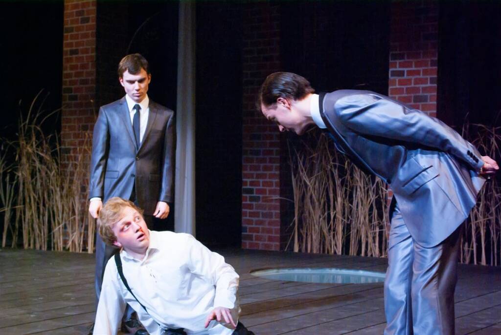 Three actors perform a dramatic scene on stage. One man in a suit is standing, another is bending forward with hands behind his back, and a third man, wearing a white shirt, is sitting on the ground. The background is dark with reeds and brick walls.