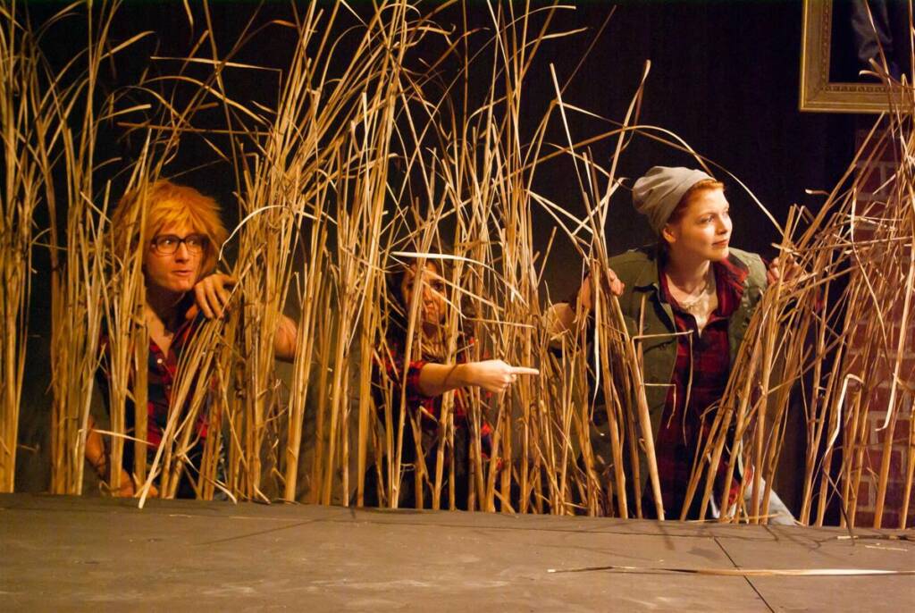 Three people in casual clothing crouch behind tall reeds on a stage, with one person pointing forward. They appear to be part of a theater performance.