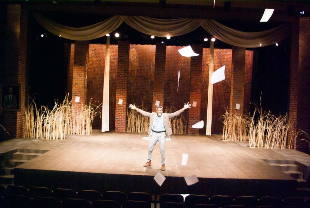 A person is standing in the center of a stage, arms outstretched, as sheets of paper fall around them. The stage is set with tall, dry grasses and illuminated against a brick backdrop. The theater is dimly lit with empty seats visible in the foreground.