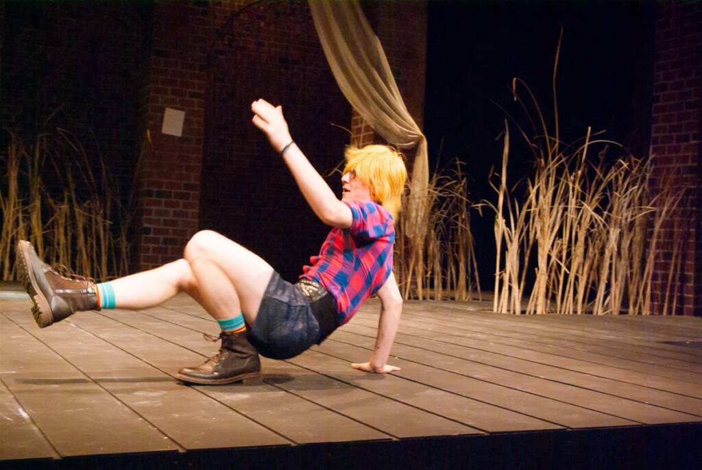 A person with short blonde hair, wearing a red and blue plaid shirt, denim shorts, and boots, performs a dynamic dance move on a wooden stage. Tall grass and brick walls are in the background.